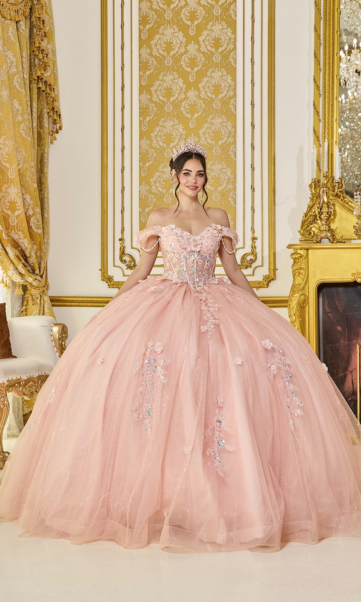 A young woman in the Quinceanera Dress 5096 By Dancing Queen in blush, featuring floral details and glitter tulle, stands smiling in an ornate gold-decorated room, wearing a tiara.
